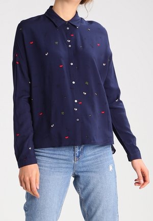 Marineblauw overhemd met knopen, bedrukt met kleine rode lippen, ogen en bladeren, gedragen met lichtblauwe gescheurde spijkerbroek door een staande persoon.