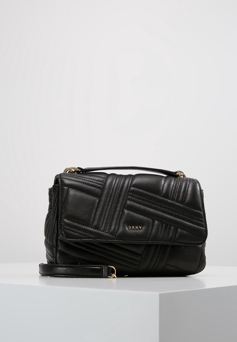 Sac à main matelassé en cuir noir DKNY avec logo doré, poignée supérieure et bandoulière amovible, posé sur un socle blanc sur fond uni.