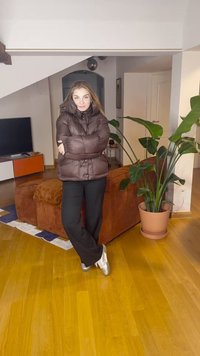 Kvinde med langt hår iført en brun dunjakke og sorte bukser, stående med armene krydset i en stue nær en stor potteplante.