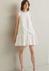 Bijela haljina bez rukava s okruglim izrezom, detaljem u obliku mašne na sredini i slojevitim volanima na rubu. Glatka tkanina, A-linijski silueta.
