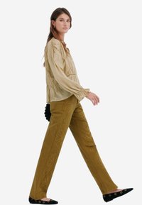 Blusa testurizzata beige chiaro con maniche a palloncino, abbinata a pantaloni giallo senape con ricami floreali e ballerine nere con decorazioni.
