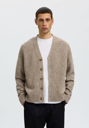 Mand med kort mørkt hår iført beige knap-cardigan over hvid skjorte og sorte bukser, stående med hænderne i lommerne mod ensfarvet baggrund.