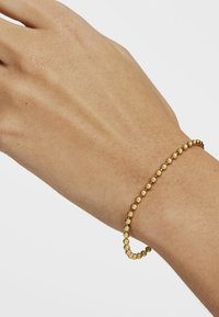 Pulsera de cuentas de oro en una muñeca, con pequeñas cuentas esféricas, superficie lisa, diseño flexible y un brillo sutil.
