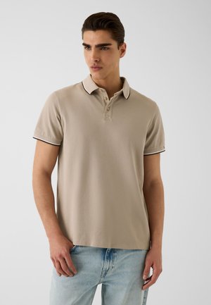 Jeune homme portant un polo beige à manches courtes avec bordure blanche et un jean bleu clair, debout devant un fond gris uni.