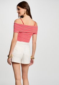 Top de rayas rojas y blancas con hombros al aire y tirantes finos, combinado con pantalones cortos blancos de cintura alta que tienen bolsillos traseros y una textura suave.