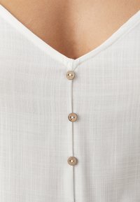 Blusa bianca con scollatura a V, caratterizzata da un tessuto testurizzato, bottoni in legno lungo la parte frontale e orlo liscio.