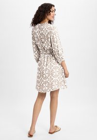 Patterned beige dress with long sleeves, a tie belt, and a relaxed silhouette. Features white decorative motifs and a knee-length cut.