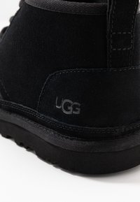 Detailní záběr na černou semišovou kotníkovou botu UGG, ukazující texturovanou podešev a švy na bílém pozadí.