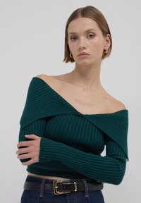 Maglione teal a coste scollato con maniche lunghe. Tessuto morbido e design aderente. Abbinato a una cintura nera sopra jeans a vita alta.