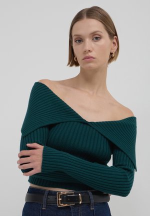 Maglione teal a coste scollato con maniche lunghe. Tessuto morbido e design aderente. Abbinato a una cintura nera sopra jeans a vita alta.