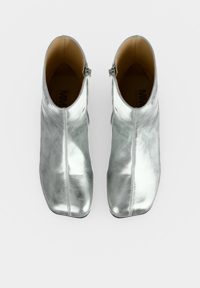 Paire de bottines argentées brillantes avec des fermetures éclair latérales, des bouts carrés arrondis et une doublure intérieure marron clair, vues de dessus sur un fond blanc.