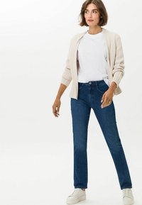 Cardigan beige a costine sopra una t-shirt bianca, abbinato a jeans blu scuro e scarpe bianche. Design semplice senza motivi o accenti visibili.