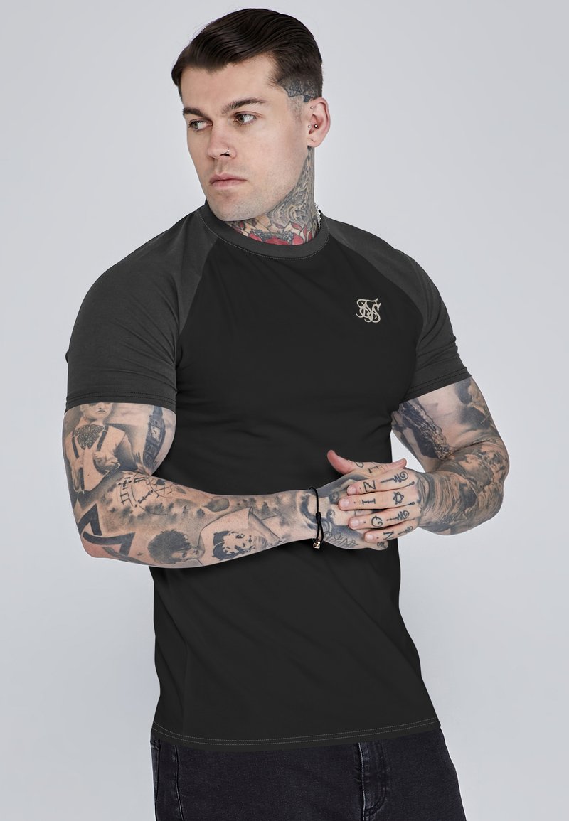 Schwarzes, eng anliegendes T-Shirt mit dunkelgrauen Raglan-Ärmeln, versehen mit einem kleinen goldenen Logo auf der Brust und einem glatten, dehnbaren Stoff.