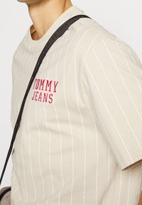 Béžové pruhované tričko s bílými vertikálními pruhy, s červeným vyšitým nápisem "TOMMY JEANS" na hrudi a kulatým výstřihem. Viditelný černý popruh.