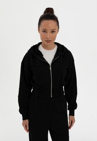 Mustat vetoketjuhupparit, joissa on nyöritettävä huppu, pehmeä kangas ja ribbatut hihansuut. Yhdistetty korkeavyötäröisten mustien housujen kanssa. Yksinkertainen, moderni muotoilu.