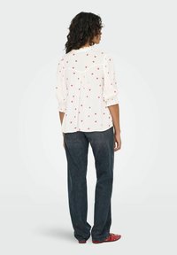 Mujer con cabello rizado, vistiendo una blusa blanca con corazones rojos, jeans oscuros holgados y zapatos rojos de punta, de pie de espaldas sobre un fondo liso.