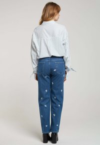 Pantalon en denim bleu avec broderie florale blanche et coupe droite, associé à une chemise à boutons rayée bleue et blanche.