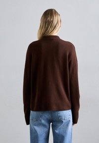 Rjav pleten pulover z rebrastim robom in manšeti, ki ima sproščen kroj. Kombinira se z svetlo modrimi kavbojkami, kar prikazuje sproščeno oblačilo s hrbtne strani.