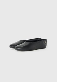Zwarte platte schoenen van soepel leer met een ronde neus en een laag profiel ontwerp. Een klein zilveren logo accent is aanwezig op de hak.