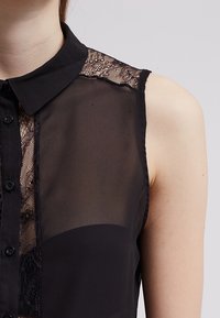 Sort ærmeløs bluse med et gennemsigtigt stof, blonde detaljer på skuldrene og en knaplukning foran. Let tekstur, tætsiddende design.