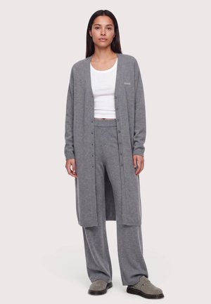 Femme portant un long cardigan gris, un pantalon large assorti, un haut court blanc et des chaussures grises, debout devant un fond uni.