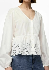 Camicia bianca in cotone con scollo a V, maniche lunghe a sbuffo, chiusura frontale con bottoni e orlo scalloppato con motivi floreali intagliati.