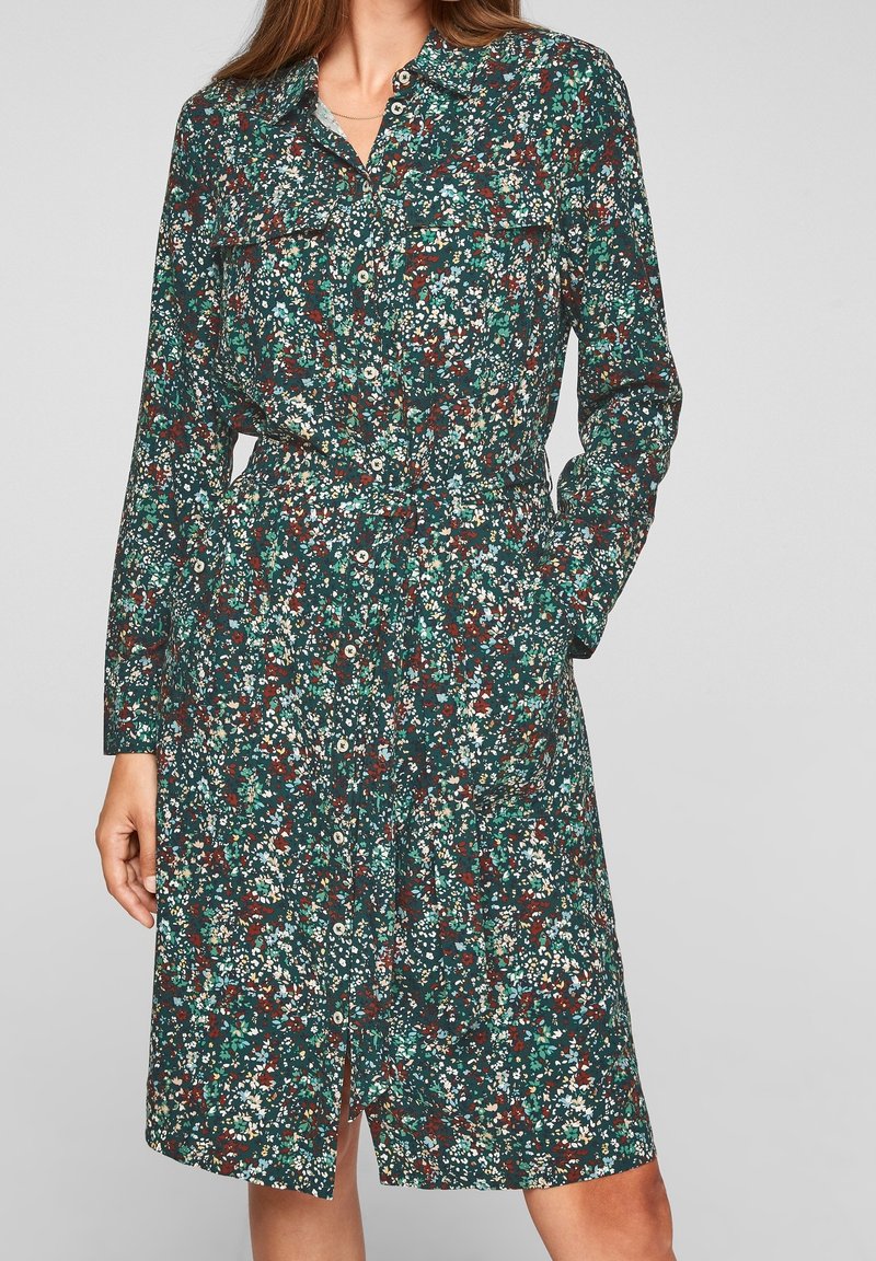 Femme portant une robe fleurie à manches longues boutonnée, aux couleurs vert, blanc et rouille, avec une ceinture à la taille, debout devant un fond uni.