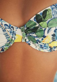 Bikini zgornji del z rožnatim potiskom, ki prikazuje zelene liste in rumene poudarke, z naborki in gladko teksturo tkanine.