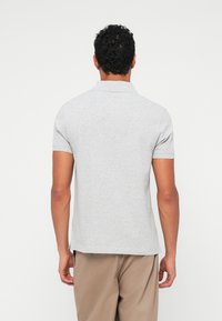 Homme de dos portant un polo gris clair à manches courtes et un pantalon beige sur un fond blanc uni.