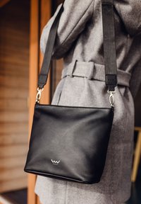 Černá kožená kabelka přes tělo s hladkou texturou, stříbrnými detaily a nastavitelným popruhem. Malé logo umístěné na přední straně.