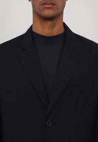 Zwarte blazer van soepel materiaal. Voorzien van een enkele knoopsluiting, geknipte revers en een borstzak. Draagt over een donkere coltrui.