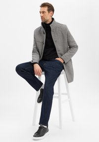 Man in een grijze jas, zwarte coltrui, donkere spijkerbroek en zwarte sneakers zittend op een witte kruk tegen een effen achtergrond.