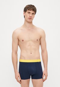 Cuecas boxer de algodão azul-marinho com uma cintura elástica amarela exibindo a palavra "HAPPY" em letras maiúsculas. Design leve e ajustado.