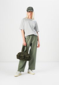 Sivé oversized tričko, zelené voľné nohavice, sivá čiapka a zelená kordurová taška s vreckami. Biele tenisky dopĺňajú outfit.