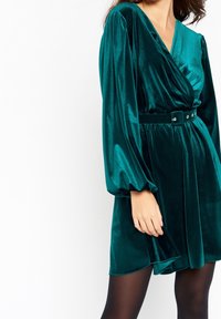 Teal sammetklänning med långa puffärmar, omslagdesign, bälte i midjan och slät textur. Har en lätt lyster och A-linjeform.