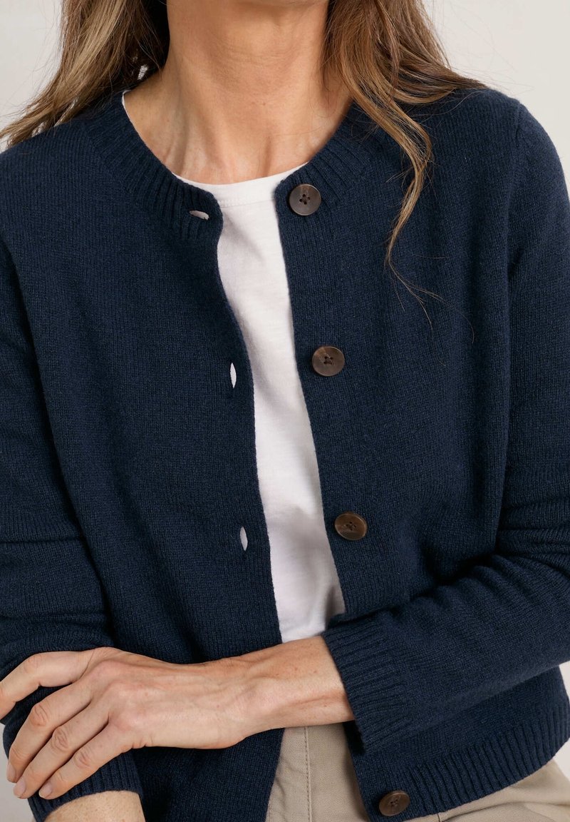Femme portant un cardigan bleu marine boutonné par-dessus une chemise blanche, les bras croisés, pantalon beige visible en bas de l'image.