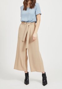 Jasnoniebieski krótki top z krótkimi rękawami, zestawiony z beżowymi szerokimi spodniami na pasku z wiązaniem, noszony z czarnymi botkami na obcasie. Gładkie tekstury.