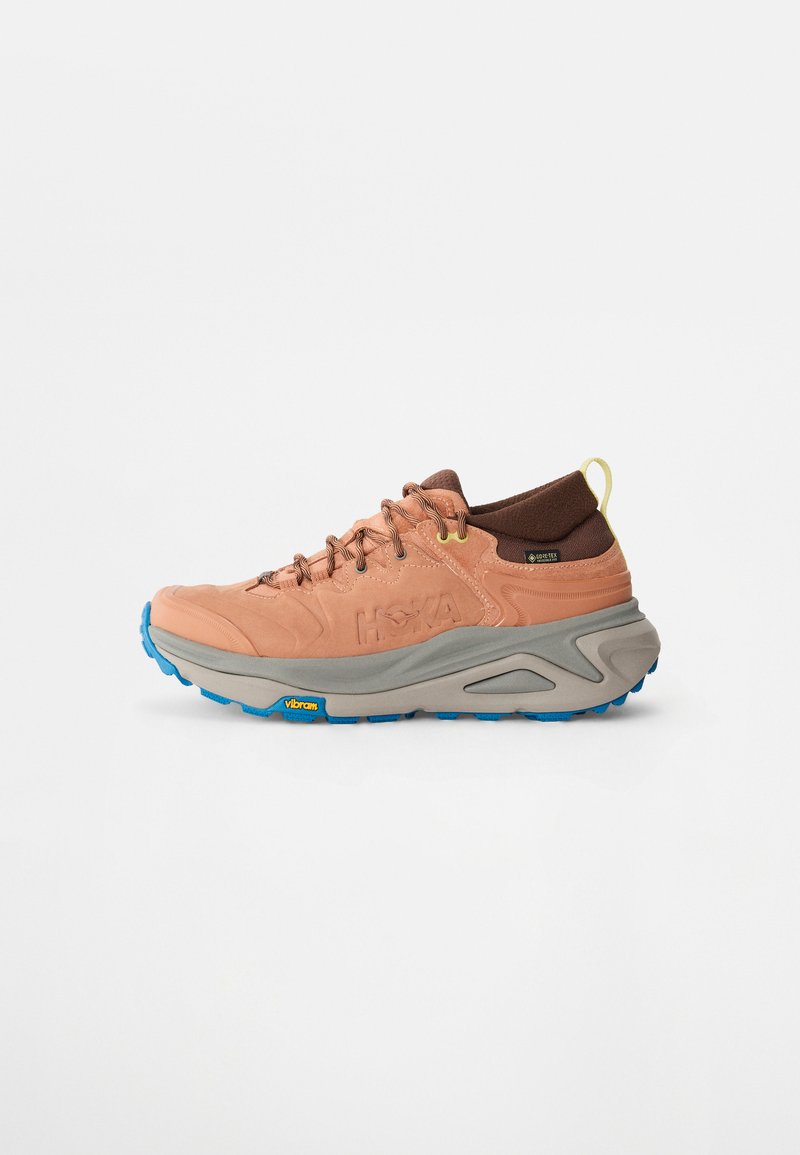 Chaussure de randonnée marron clair avec semelle intermédiaire grise et semelle extérieure bleue. Dispose de lacets, d'une tige texturée et d'une languette de talon renforcée avec logo. Marque Vibram visible.
