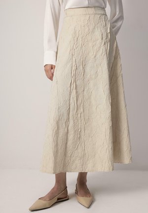 Cremefarbener, langer, zerknitterter Rock aus strukturiertem Stoff mit hoher Taille und fließendem Design. Getragen mit spitzen, nude-farbenen Ballerinas.