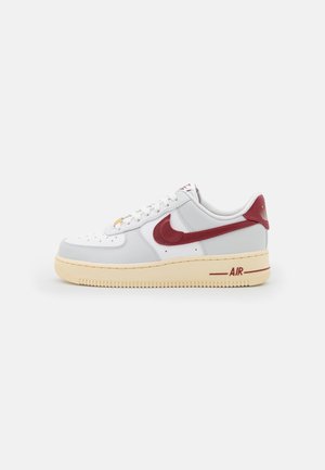 Zapatilla baja blanca con suela beige, logo rojo de Nike y lengüeta roja en el talón, mostrada de lado sobre un fondo liso.