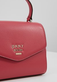 Punainen tekstiilinäyte DKNY -laukkua, jossa on yläkahva ja kultainen logo "DKNY NEW YORK EST. 1989" etuosion läpällä, harmaata taustaa vasten.