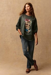 Sweatshirt vert foncé avec un portrait floral de Frida Kahlo, associé à un jean bleu délavé et des chaussures slip-on marron.