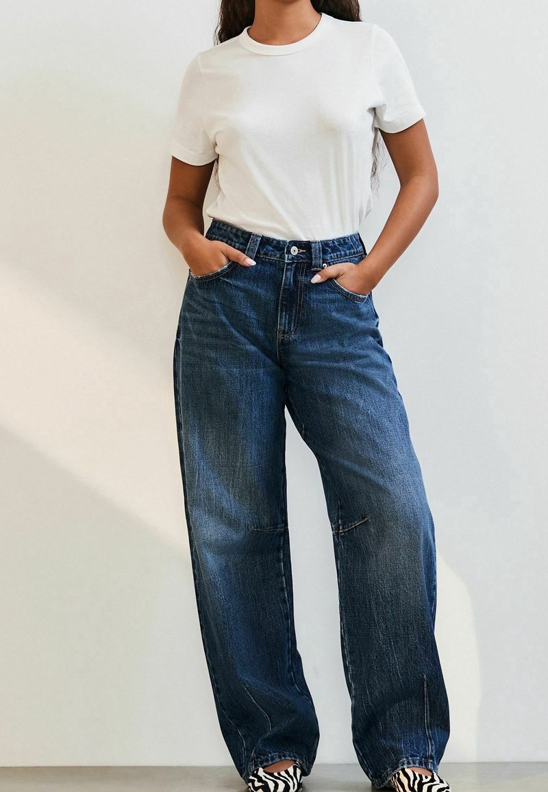 Femme portant un t-shirt blanc à manches courtes, jean bleu taille haute à jambes larges, chaussures à imprimé zèbre, debout les mains dans les poches.