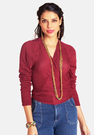 Frau trägt ein langärmeliges Oberteil in Kastanienbraun, blaue Jeans, eine bunte Perlenkette, passende lange Ohrringe und ein mehrfarbiges Armband, steht mit heruntergelassenen Händen.