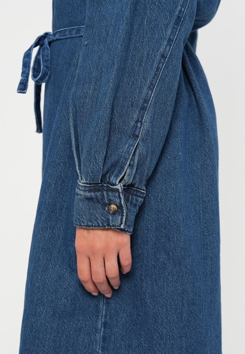 Dżinsowa sukienka z dopasowaną talią, długimi bufkowymi rękawami i zapięciem na guziki przy mankietach. Posiada tonowany pasek w talii oraz intensywny niebieski kolor z teksturowanej tkaniny.