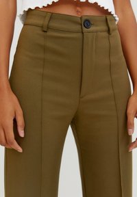 Close-up van een persoon die hooghangende olijfgroene broek draagt met een zwarte knopen en een cropped witte top, handen ontspannen aan de zijkanten.