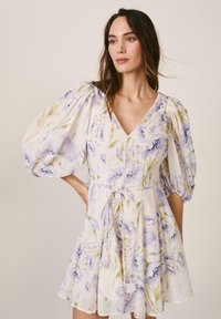 Mujer con un vestido blanco floral de mangas abullonadas y escote en V, atado a la cintura, posando frente a un fondo liso.