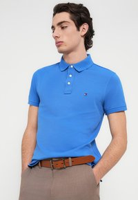 Μπλε βαμβακερό πόλο πουκάμισο με γιακά, πλάκα με τρία κουμπιά και μικρό λογότυπο εμπορικού σήματος στο στήθος. Καφέ ζώνη με μεταλλική αγκράφα.