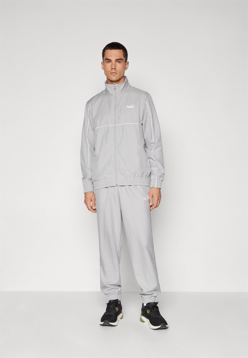 Lys grå træningsdragt med zip-jakke og elastiske manchetter, prydet med hvide accentstriber og Puma-logoet. Båret med sorte sportsko.