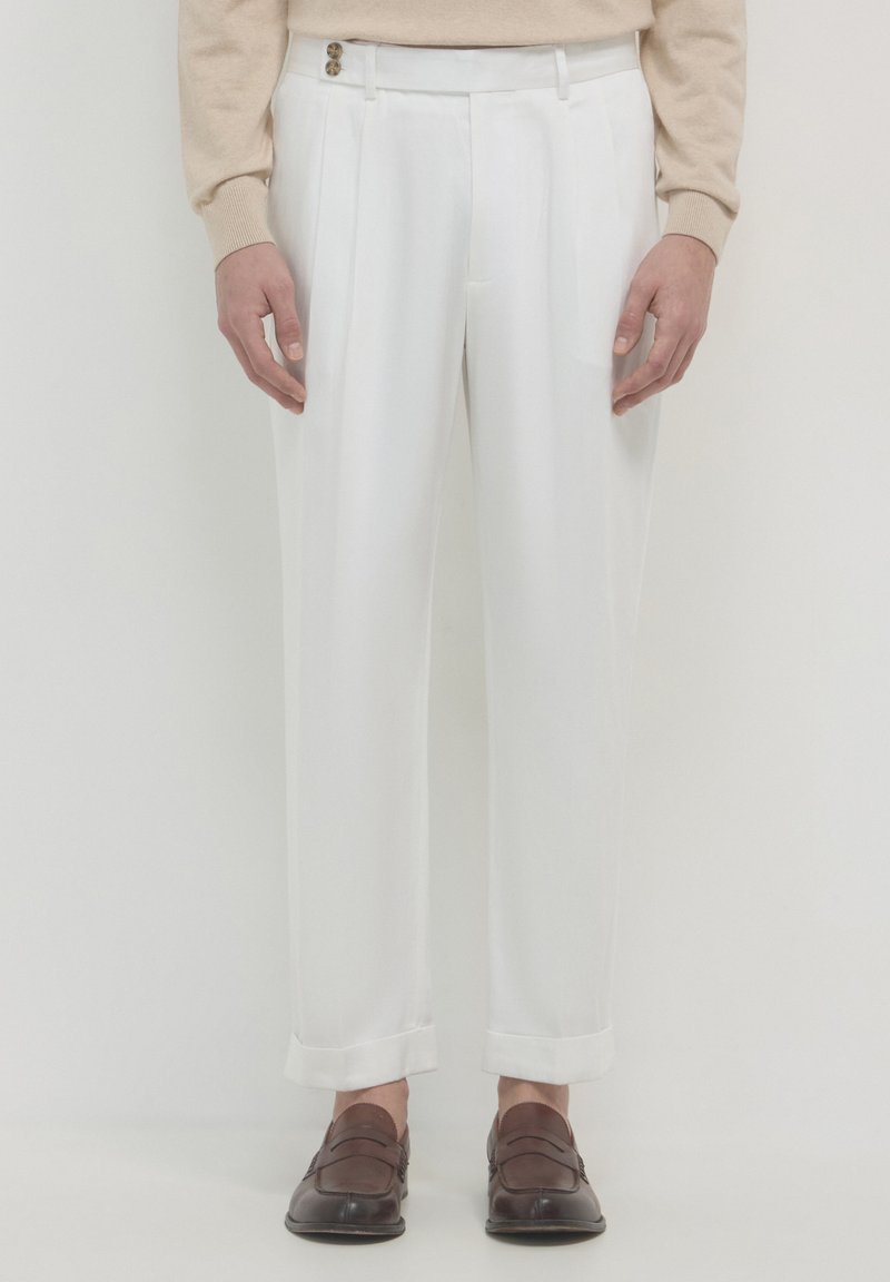 Pantaloni bianchi plissettati con una vestibilità sartoriale, dotati di un frontale piatto, orli con risvolto e due bottoni in vita. Indossati con mocassini marroni.